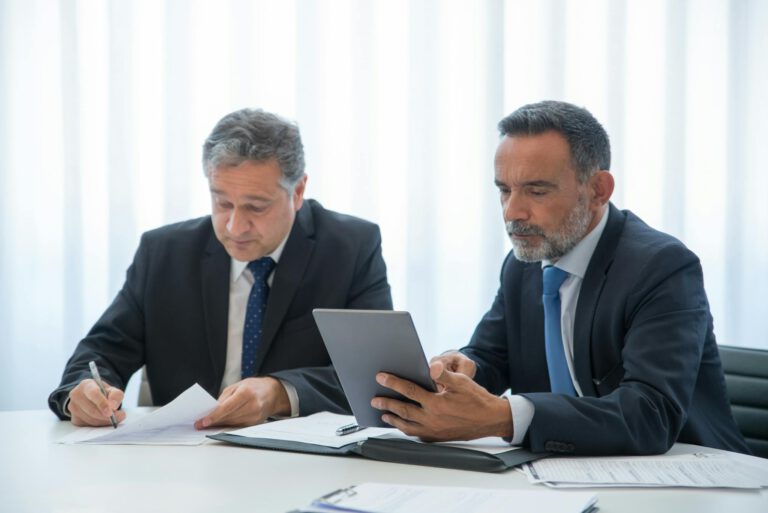 Two businessmen in a modern office reviewing documents and working on a tablet.